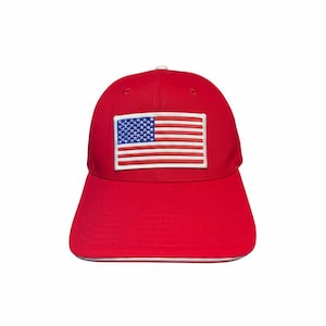 Op de afbeelding: Rode baseballcap met een witte rand en een rechthoekige patch met de Amerikaanse vlag. De vlag heeft een blauw veld met witte sterren en rode en witte strepen.
