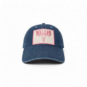 Op de afbeelding: Een blauwe denim baseball cap met een gebogen klep. De pet heeft een rechthoekige patch met het woord "WALLEN" in roze letters en een roze bull skull graphic.