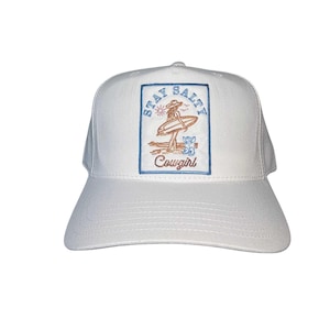 May include: White baseball cap with a rectangular patch featuring a surfer girl graphic and the words "Stay Salty Cowgirl" in blue and orange. The cap has a curved peak and a structured crown.