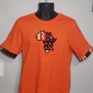 Peut inclure: T-shirt orange avec un motif coloré du continent africain sur le devant. Les manches ont des poignets à motifs. Le motif présente des accents rouges, jaunes et bruns. Le t-shirt est présenté sur un mannequin noir.
