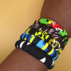 May include: A stack of fabric-covered bracelets in various colors and patterns. The bracelets include black with white accents, blue and yellow stripes, yellow with black and white designs, and red, green, and black patterns.