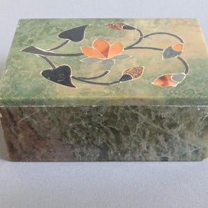 May include: A rectangular, green marble box with a floral inlay design. The lid features a central orange flower with black and brown accents. The box has a marbled texture and a simple, elegant design.