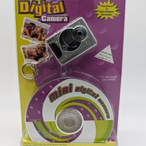 May include: A silver Smart Mini Digital Camera keychain in purple and yellow retail packaging. The package includes a circular disc, text reading Smart Mini Digital Camera, and system requirements for Windows 98, ME, 2000, and XP. The camera has a lens.