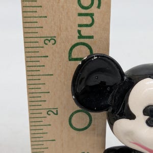 May include: A close-up of a vintage Mickey Mouse figurine with black ears and a white face. A wooden ruler is next to the figurine. The figurine has a glossy finish. The ruler shows the numbers 2 and 3.