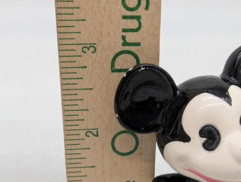 May include: A close-up of a vintage Mickey Mouse figurine with black ears and a white face. A wooden ruler is next to the figurine. The figurine has a glossy finish. The ruler shows the numbers 2 and 3.