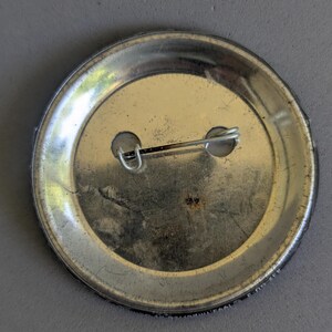 Op de afbeelding: Zilverkleurige metalen knooprug met een speld en twee gaten. De knoop heeft een cirkelvormige vorm met een licht versleten uiterlijk. De speld is met een kleine sluiting aan de achterkant bevestigd.
