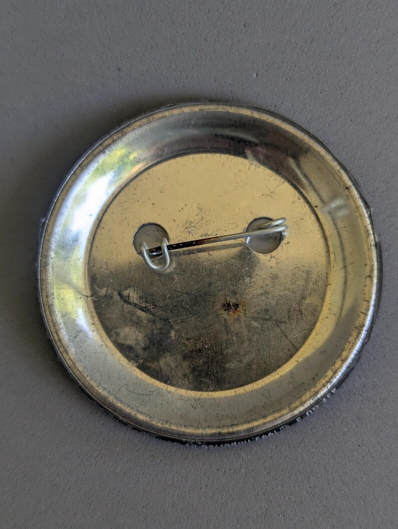 Op de afbeelding: Zilverkleurige metalen knooprug met een speld en twee gaten. De knoop heeft een cirkelvormige vorm met een licht versleten uiterlijk. De speld is met een kleine sluiting aan de achterkant bevestigd.