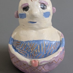 May include: A handmade ceramic sculpture of a stylized figure. The figure has a white face with blue and purple accents, including a blue stripe across the chest and a purple patterned base. The sculpture has a unique, artistic design.