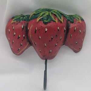 May include: A decorative wall hook featuring three red strawberries with green leaves. The strawberries have white speckles and a black metal hook extending from the bottom. The hook is designed to hang items.