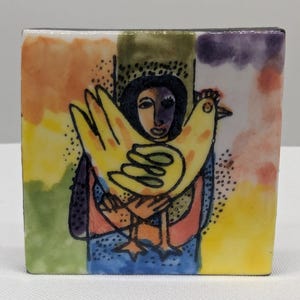 May include: A square ceramic tile featuring a folk art illustration. The artwork depicts a person holding a large yellow chicken. The background has colorful abstract shapes in orange, green, and purple. The person is wearing a blue top and a black head covering.