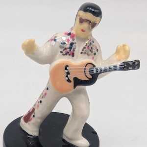 May include: A ceramic figurine of a musician in a white jumpsuit with colorful embellishments, playing a guitar. The figurine has sunglasses and a black hairstyle. The base is black.