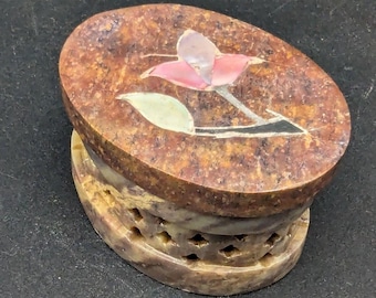 May include: Oval-shaped, brown stone box with a carved base. The lid features a pink and white flower inlay. The box has a decorative, carved design around the base, adding a touch of elegance. The box is a decorative item.