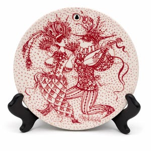 May include: A round, off-white decorative plate with a red illustration of two figures in ornate costumes, one playing a lute. The plate has a small hole at the top and is displayed on a black stand. The design features a floral pattern.