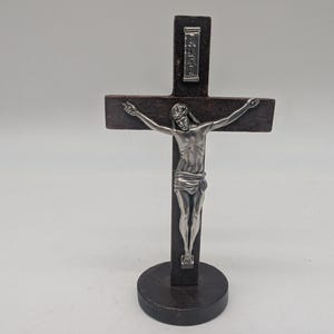 May include: A dark brown wooden crucifix with a silver figure of Jesus Christ. The cross has a rectangular shape with a small plaque at the top. The figure is detailed with outstretched arms and a draped loincloth, mounted on a round base.
