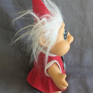 Puede incluir: Mu&ntilde;eca troll vintage con un vestido rojo y un gorro puntiagudo a juego. La mu&ntilde;eca tiene pelo blanco y esponjoso y ojos azules. El gorro est&aacute; rematado con un pomp&oacute;n blanco. La piel de la mu&ntilde;eca es de color canela claro.