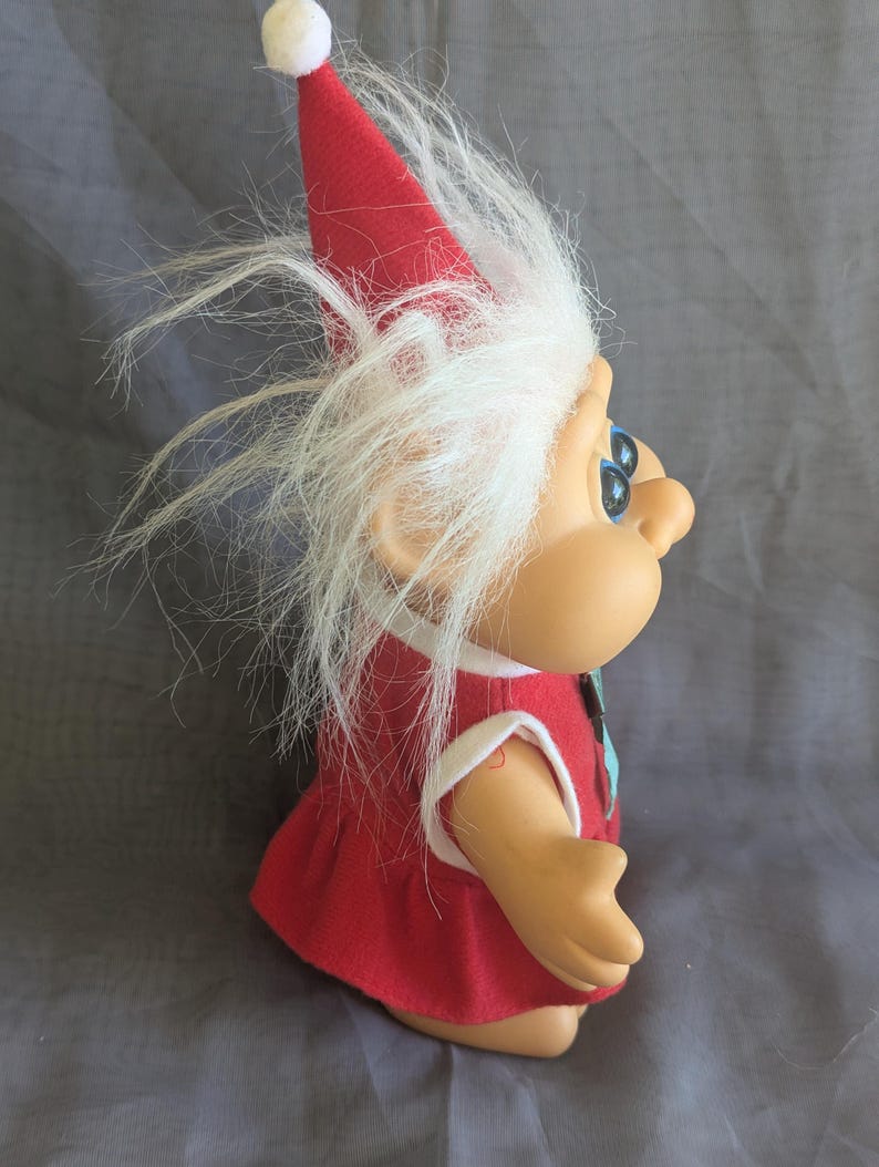 Puede incluir: Mu&ntilde;eca troll vintage con un vestido rojo y un gorro puntiagudo a juego. La mu&ntilde;eca tiene pelo blanco y esponjoso y ojos azules. El gorro est&aacute; rematado con un pomp&oacute;n blanco. La piel de la mu&ntilde;eca es de color canela claro.