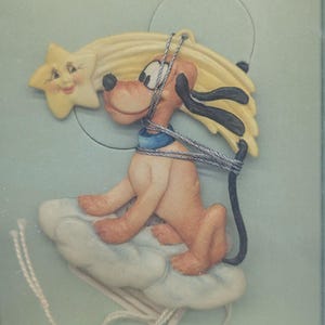 May include: A decorative ornament featuring a cartoon dog character sitting on a cloud, with a shooting star and a smiling star. The dog is light brown with black ears and a blue collar. The ornament has a string for hanging.