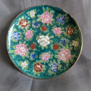 May include: A small, round decorative plate with a teal background and a floral pattern. The flowers are in shades of pink, white, red, and blue, with green leaves. The plate has a gold rim.