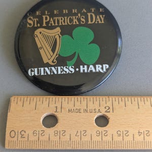 Op de afbeelding: Een ronde zwarte St. Patrick's Day-knop met gouden tekst die luidt "CELEBRATE ST. PATRICK'S DAY" en "GUINNESS HARP". De knop heeft een gouden harp en een groene klaver. Een houten liniaal ligt eronder.
