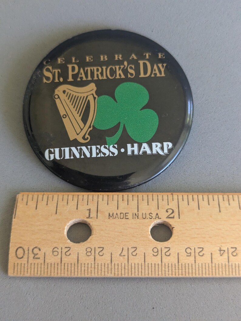 Op de afbeelding: Een ronde zwarte St. Patrick's Day-knop met gouden tekst die luidt "CELEBRATE ST. PATRICK'S DAY" en "GUINNESS HARP". De knop heeft een gouden harp en een groene klaver. Een houten liniaal ligt eronder.