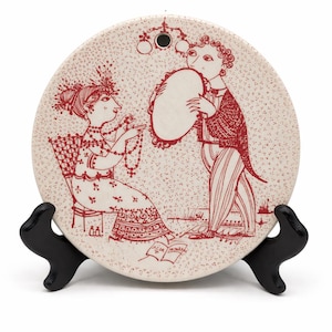 May include: A round, off-white decorative plate with a red illustration of two figures. One figure is seated, adorned with jewelry, while the other holds a tambourine. The plate is displayed on a black stand.