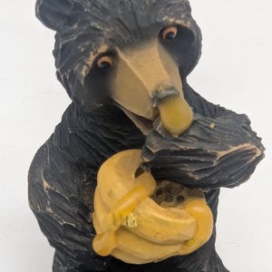 May include: A carved wooden bear figurine in black and tan, holding a yellow honey pot. The bear has detailed fur texture and is eating honey from the pot. The figurine is a decorative item.
