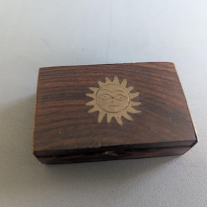 May include: A rectangular dark brown wooden box featuring a brass inlay of a stylized sun face on the lid. The wood grain is visible across the surface of the lid, and the box has a simple, flat construction with straight edges and corners.