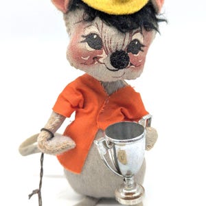 May include: A vintage stuffed mouse doll wearing an orange jacket and a yellow cap. The mouse holds a silver trophy and a string. The doll has black hair and rosy cheeks. The doll is a collectible item.