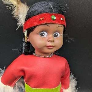 May include: A vintage doll with dark hair and a red headband adorned with beads and a green jewel. The doll wears a red dress with a green belt and white fur trim. A feather is attached to the doll's head.