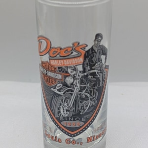 May include: Clear shot glass with the text "Doc's Harley-Davidson" in orange and black. The glass features a motorcycle graphic and the words "Since 1955" and "Louis Co., Misso".