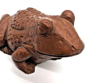 May include: A detailed, brown wooden frog sculpture. The frog is carved with textured skin and closed eyes, giving it a lifelike appearance. The sculpture is set against a white background, highlighting the frog's form.
