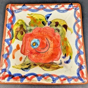 Peut inclure: Petit plat carré en céramique avec un motif floral peint à la main. Au centre, une rose rouge entourée de feuilles vertes et jaunes. Le bord présente un motif ondulé rouge et bleu sur fond blanc. Bordure marron.