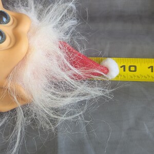 Puede incluir: Primer plano de la cara de una mu&ntilde;eca con ojos azules, tez bronceada y barba blanca. Se ve un gorro de Pap&aacute; Noel rojo con un pomp&oacute;n blanco. Una regla amarilla muestra que el gorro mide unos 2,5 cm.