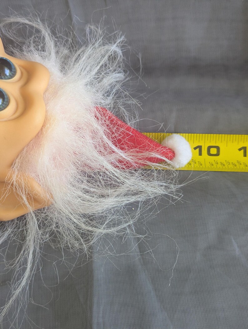 Puede incluir: Primer plano de la cara de una mu&ntilde;eca con ojos azules, tez bronceada y barba blanca. Se ve un gorro de Pap&aacute; Noel rojo con un pomp&oacute;n blanco. Una regla amarilla muestra que el gorro mide unos 2,5 cm.