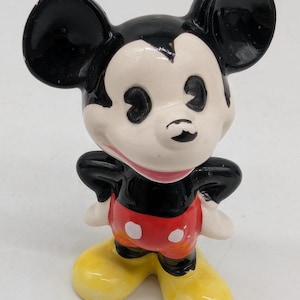 May include: A ceramic Mickey Mouse figurine. The figure features black ears, a black torso, and red shorts with white buttons. The face is white with black eyes and a small black nose. The shoes are yellow, and the figure is posed with hands on hips.