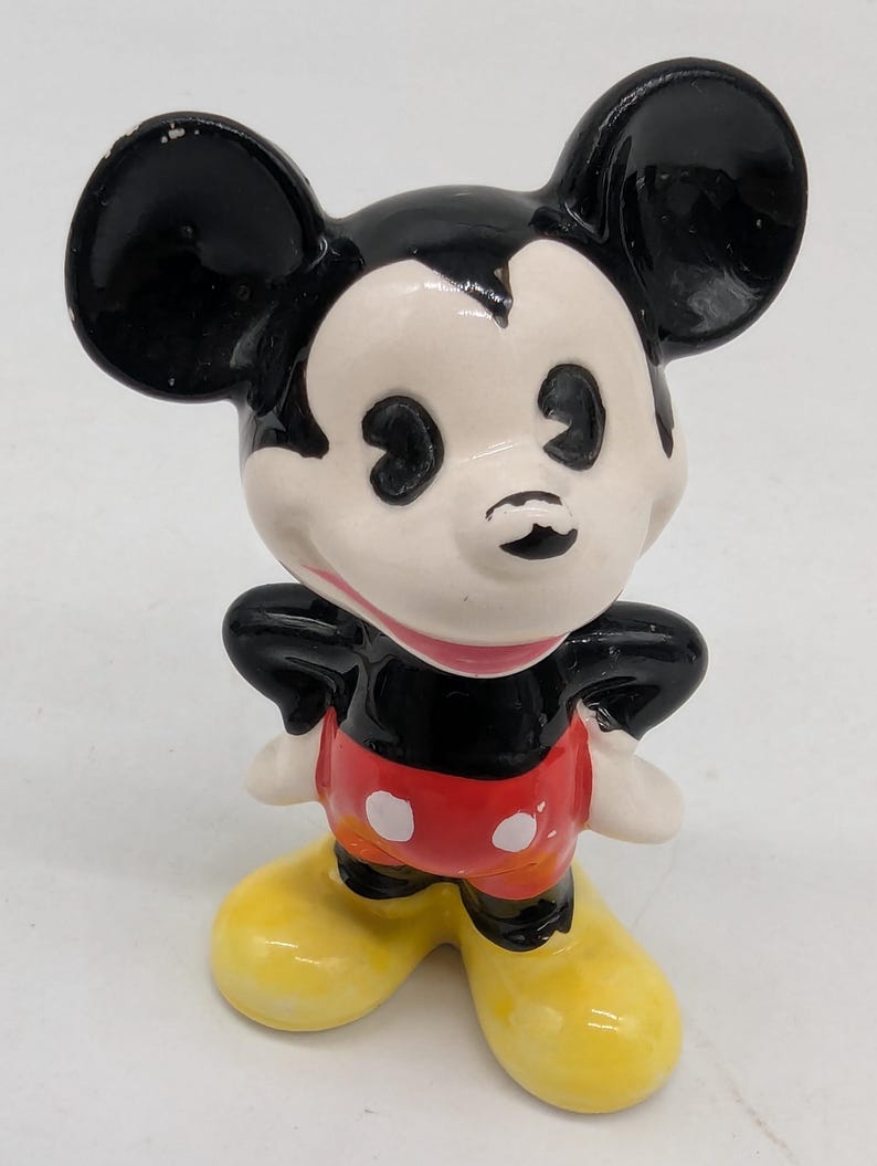 May include: A ceramic Mickey Mouse figurine. The figure features black ears, a black torso, and red shorts with white buttons. The face is white with black eyes and a small black nose. The shoes are yellow, and the figure is posed with hands on hips.