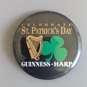 Op de afbeelding: Zwarte ronde knop met gouden tekst "CELEBRATE ST. PATRICK'S DAY" en "GUINNESS HARP". Voorzien van een gouden harp en een groene klaver.