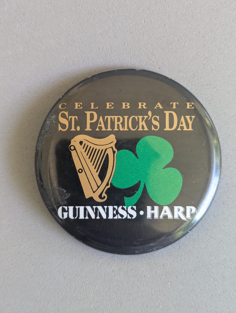Op de afbeelding: Zwarte ronde knop met gouden tekst "CELEBRATE ST. PATRICK'S DAY" en "GUINNESS HARP". Voorzien van een gouden harp en een groene klaver.