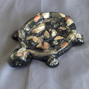 May include: A decorative turtle-shaped trinket dish. The dish is black with a mosaic of colorful shells and stones embedded in the surface. The turtle has a glossy finish and is resting on a gray surface.