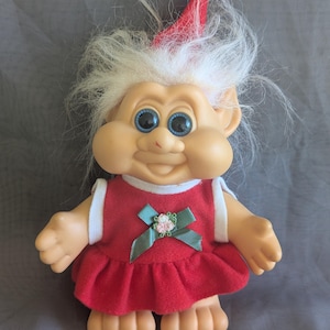 Puede incluir: Una mu&ntilde;eca troll vintage con cabello blanco y ojos azules. La mu&ntilde;eca viste un vestido rojo con ribete blanco y un lazo verde con una flor rosa. Lleva un gorro de Pap&aacute; Noel rojo y blanco.