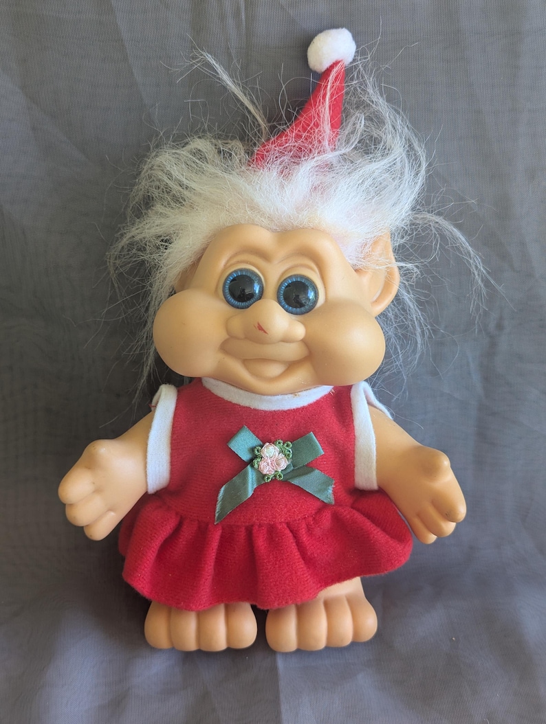 Puede incluir: Una mu&ntilde;eca troll vintage con cabello blanco y ojos azules. La mu&ntilde;eca viste un vestido rojo con ribete blanco y un lazo verde con una flor rosa. Lleva un gorro de Pap&aacute; Noel rojo y blanco.