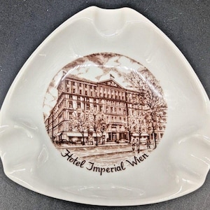 May include: White ceramic ashtray with a triangular shape and three curved indentations. The center features a brown illustration of a building with the text "Hotel Imperial Wien". The ashtray has a glossy finish.