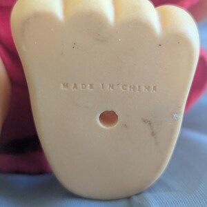 Puede incluir: Primer plano de la parte inferior de un pie de pl&aacute;stico beige claro, con las palabras "MADE IN CHINA" estampadas. El pie tiene forma redondeada con tres dedos y un peque&ntilde;o agujero. Se ve una tela roja en el fondo.