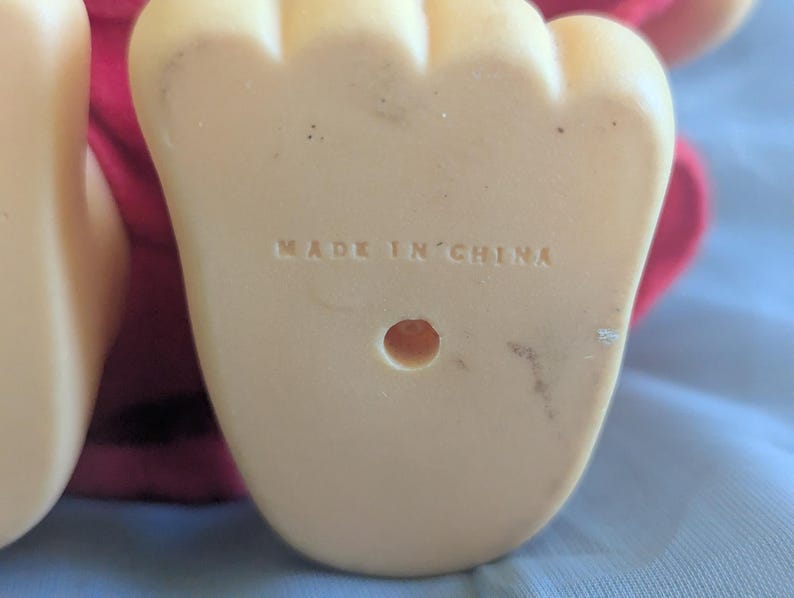 Puede incluir: Primer plano de la parte inferior de un pie de pl&aacute;stico beige claro, con las palabras "MADE IN CHINA" estampadas. El pie tiene forma redondeada con tres dedos y un peque&ntilde;o agujero. Se ve una tela roja en el fondo.