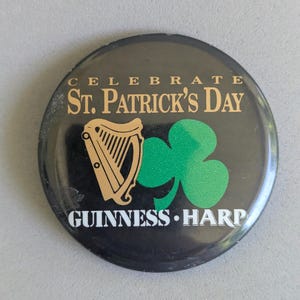 Op de afbeelding: Een ronde zwarte knoop met gouden tekst met de tekst "CELEBRATE ST. PATRICK'S DAY". Het heeft een gouden harp en een groene klaver. De tekst "GUINNESS HARP" is ook aanwezig op de knop.