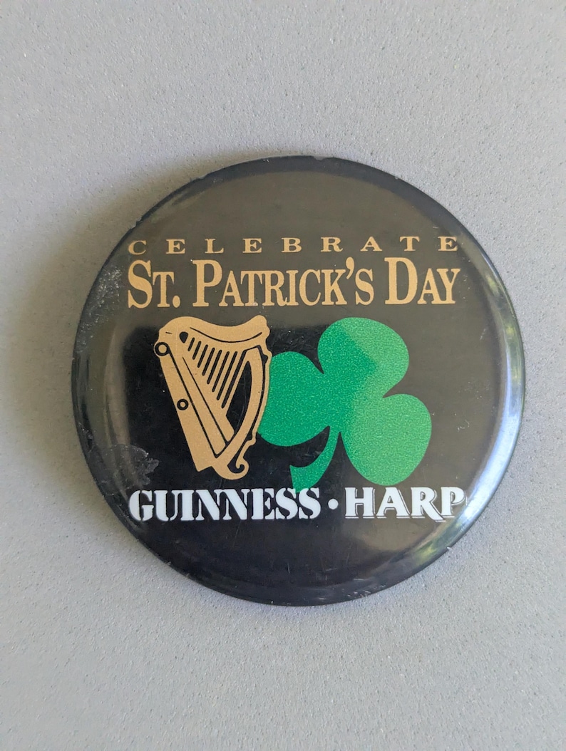 Op de afbeelding: Een ronde zwarte knoop met gouden tekst met de tekst "CELEBRATE ST. PATRICK'S DAY". Het heeft een gouden harp en een groene klaver. De tekst "GUINNESS HARP" is ook aanwezig op de knop.