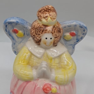 May include: A ceramic angel figurine with a smiling face, brown hair, and blue wings. The angel wears a yellow dress and a pink skirt. The wings have yellow and red accents. The hands are clasped in prayer.
