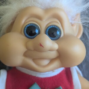 Puede incluir: Una mu&ntilde;eca Troll vintage con una expresi&oacute;n sonriente, ojos azules grandes y cabello blanco. La mu&ntilde;eca lleva un vestido rojo con ribete blanco y un lazo verde con acentos florales rosas. La cara de la mu&ntilde;eca tiene una peque&ntilde;a marca roja.