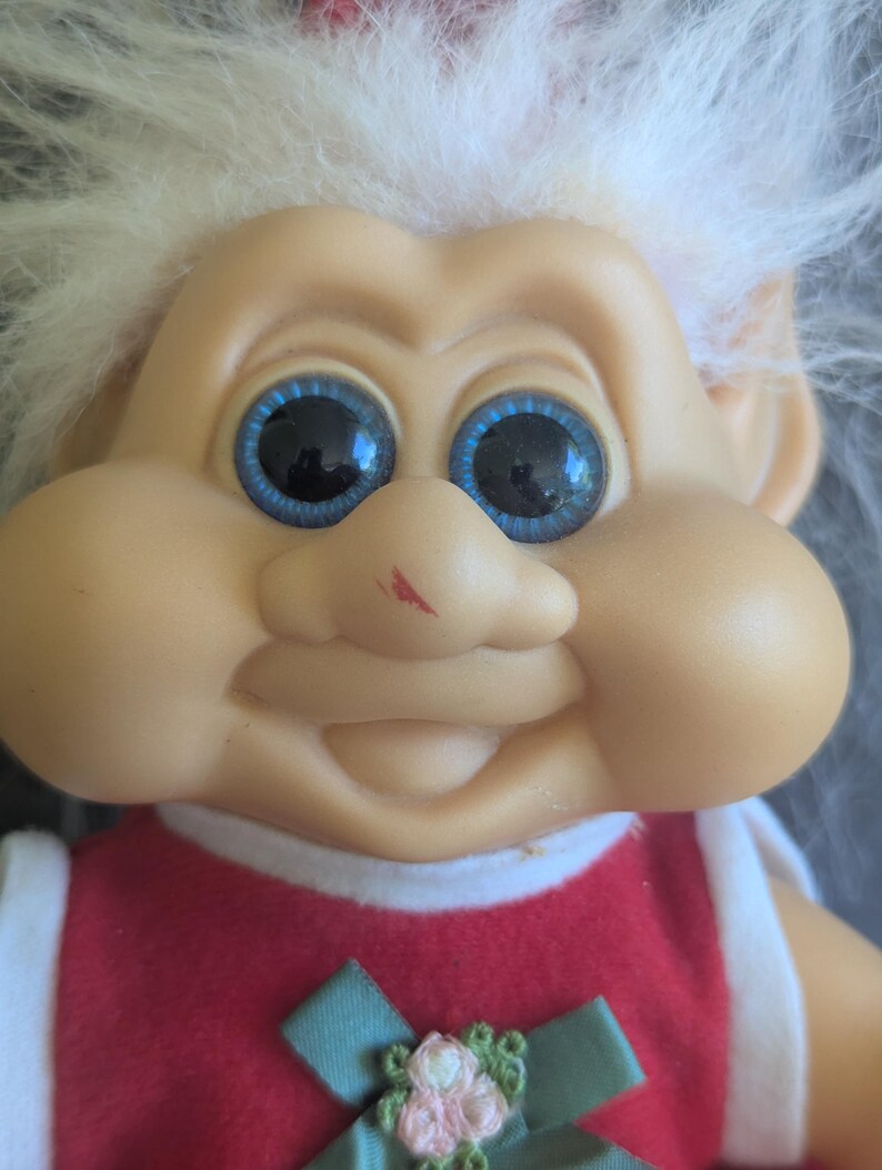 Puede incluir: Una mu&ntilde;eca Troll vintage con una expresi&oacute;n sonriente, ojos azules grandes y cabello blanco. La mu&ntilde;eca lleva un vestido rojo con ribete blanco y un lazo verde con acentos florales rosas. La cara de la mu&ntilde;eca tiene una peque&ntilde;a marca roja.