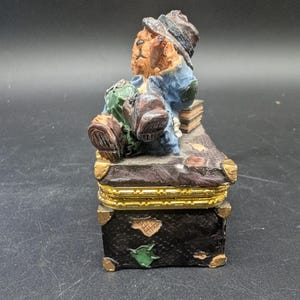 May include: A decorative trinket box featuring a seated bear figure in a hat and blue jacket, resting on a miniature trunk. The box is black with gold accents and a green leaf design.
